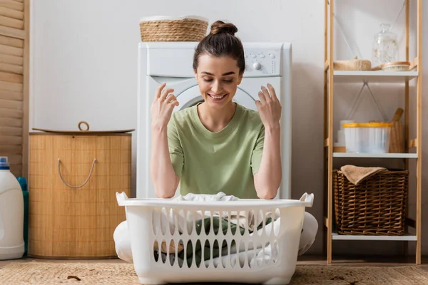 Positive brunette woman looking at basket with clothes in laundry room - foto de stock
