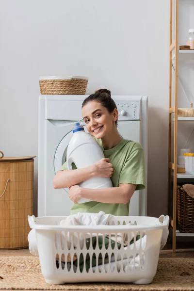 Positive woman holding washing liquid and looking at camera in laundry room - foto de stock