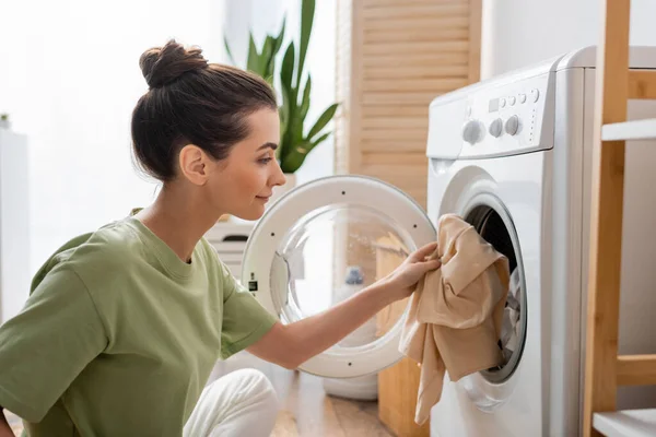 Vista lateral de la mujer joven poniendo ropa en la lavadora en casa - foto de stock