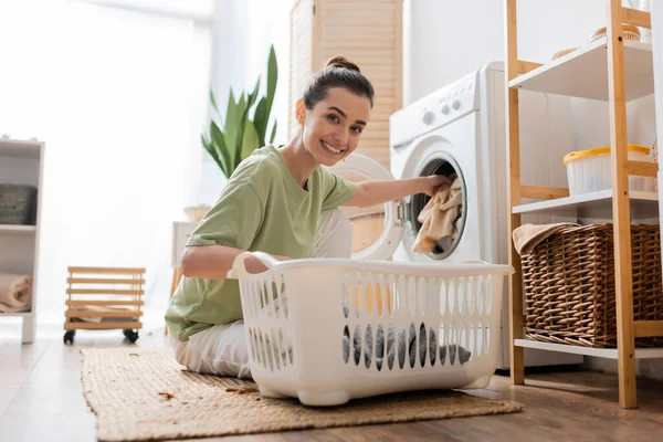 Молодая женщина улыбается в камеру, кладя одежду в стиральную машину в прачечной — стоковое фото