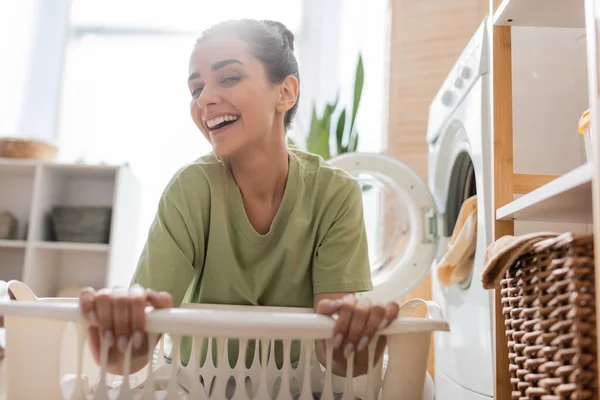 Cheerful woman looking at camera near blurred basket and washing machine at home — Fotografia de Stock