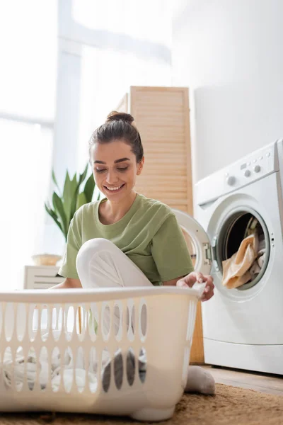 Mulher bonita sorrindo enquanto olha para a cesta com roupas perto da máquina de lavar roupa em casa — Fotografia de Stock