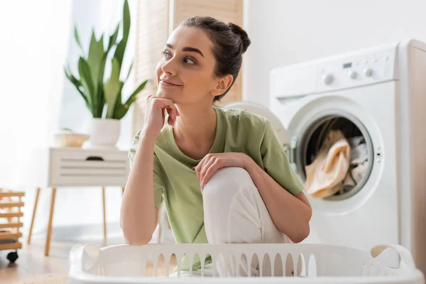 Verträumte Frau sitzt neben Korb und verschwommener Waschmaschine zu Hause — Stockfoto