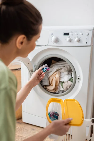 Blurred young woman holding washing capsule near machine in laundry room - foto de stock
