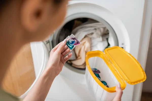 Vista cortada da mulher desfocada segurando a cápsula de lavagem e caixa perto da máquina na lavanderia — Fotografia de Stock