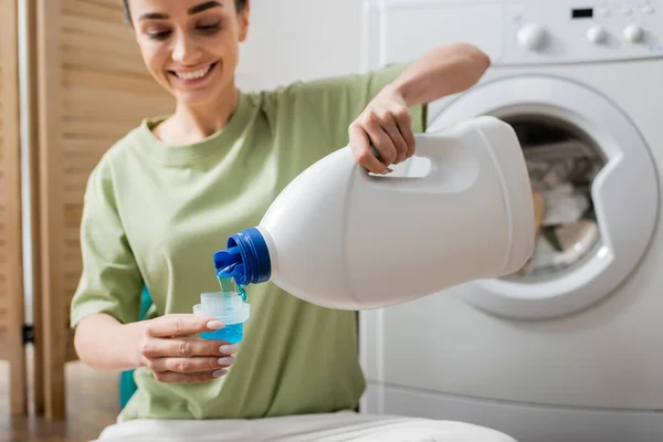 Blurred smiling woman pouring washing liquid in cap near machine in laundry room — Stock Photo
