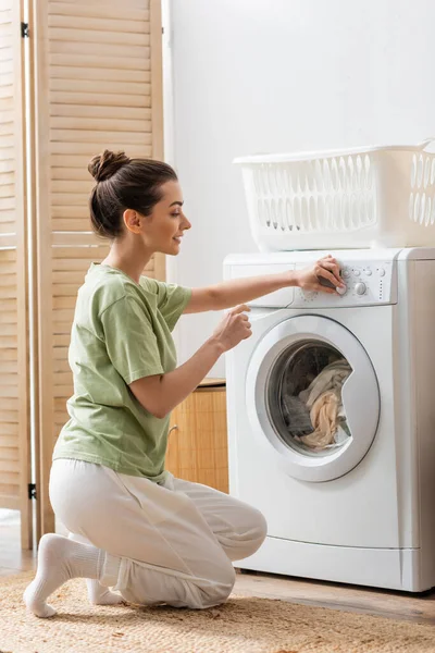 Seitenansicht einer lächelnden Frau, die zu Hause neben dem Korb die Waschmaschine schaltet — Stockfoto