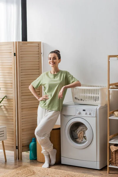 Smiling woman looking at camera while standing near washing machine in laundry room — Foto stock