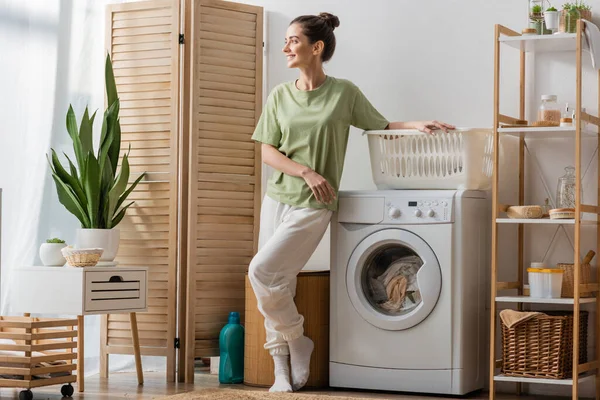 Smiling woman standing near basket and washing machine at home — Stockfoto