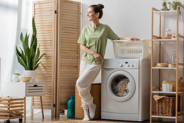 Smiling woman standing near basket and washing machine at home — Stock Photo