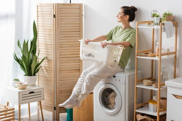 Side view of young woman holding basket while sitting on washing machine in laundry room — Stock Photo