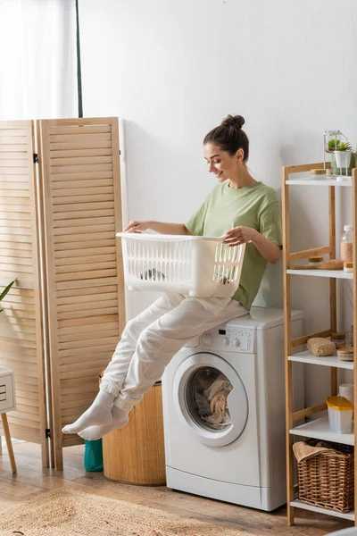 Young woman looking at basket while sitting on washing machine in laundry room - foto de stock