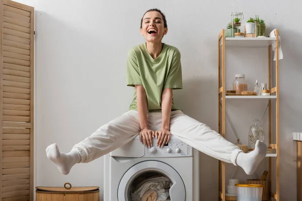 Excited woman sitting on washing machine with clothes at home - foto de stock