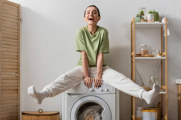Femme excitée assise sur une machine à laver avec des vêtements à la maison — Photo de stock