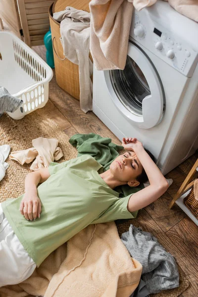 Vue du dessus de la femme fatiguée couchée autour des vêtements près de la machine à laver dans la buanderie — Photo de stock