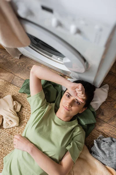 Top view of tensed woman lying on clothes near white washing machine at home — Stock Photo