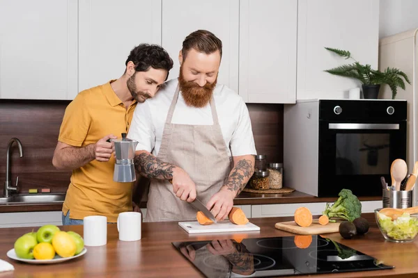 Gay homem segurando café pote perto marido corte doce batata no cozinha — Fotografia de Stock