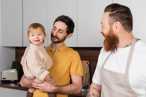 Gay parent holding smiling toddler daughter near husband in kitchen — Stock Photo
