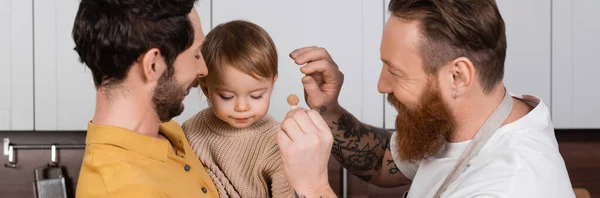 Smiling gay fathers holding barrette near daughter in kitchen, banner - foto de stock