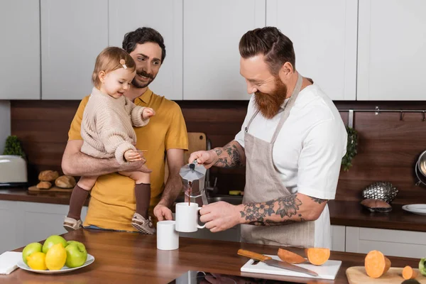Позитивний татуйований гей чоловік виливає каву біля партнера, тримаючи дочку на кухні — стокове фото