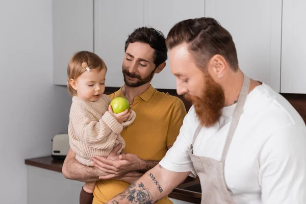 Усміхнений гей-чоловік тримає дочку з яблуком біля бородатого партнера на кухні — стокове фото
