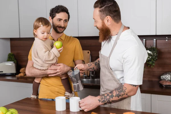 Позитивний гей-чоловік тримає гейзер кавоварку біля партнера з дочкою на кухні — стокове фото