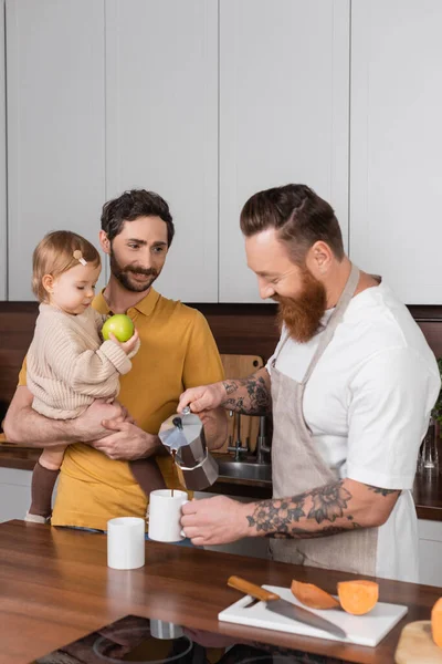 Smiling gay man holding toddler daughter with apple while partner pouring coffee in kitchen — Foto stock