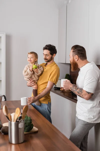 Gay couple holding coffee cups near smiling daughter with apple in kitchen — стоковое фото