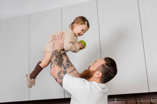 Tattooed man holding toddler daughter with apple in kitchen — Stock Photo
