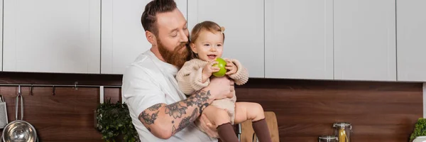 Бородатий батько тримає дівчинку-малятко з яблуком на кухні, банер — стокове фото