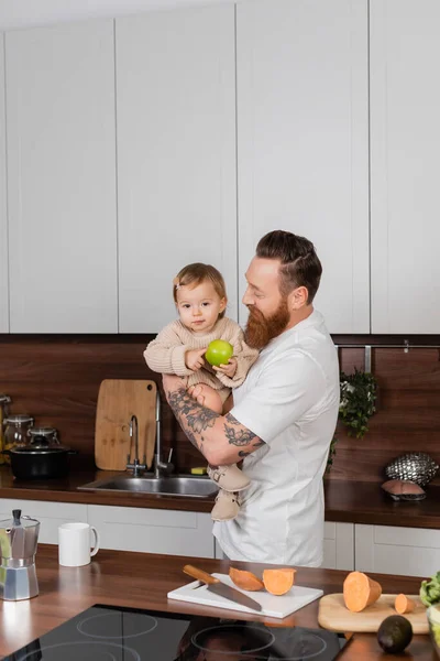 Бородатый мужчина держит маленькую дочь с яблоком на кухне дома — стоковое фото