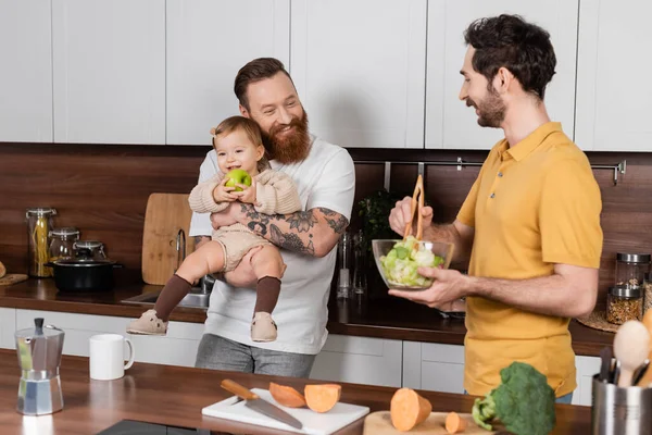 Усміхнений гей-чоловік обіймає дочку з яблуком, коли партнер готує салат на кухні — стокове фото