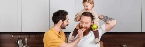 Gay uomo alimentazione marito vicino sorridente figlia con verde mela in cucina banner — Foto stock