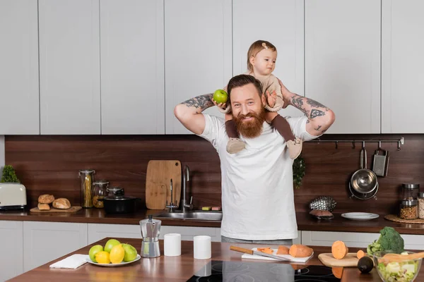 Sorrindo tatuado homem segurando criança filha com maçã perto de legumes na cozinha — Fotografia de Stock