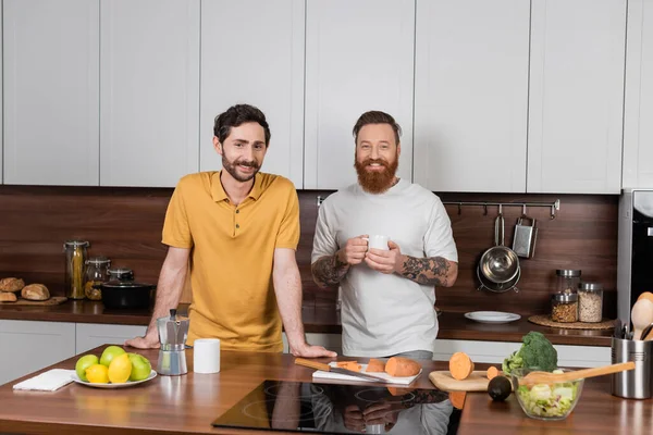 Улыбающаяся гей-пара с кофе, смотрящая в камеру на современной кухне — стоковое фото