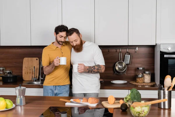 Pareja gay sosteniendo tazas de café cerca de verduras en la cocina - foto de stock