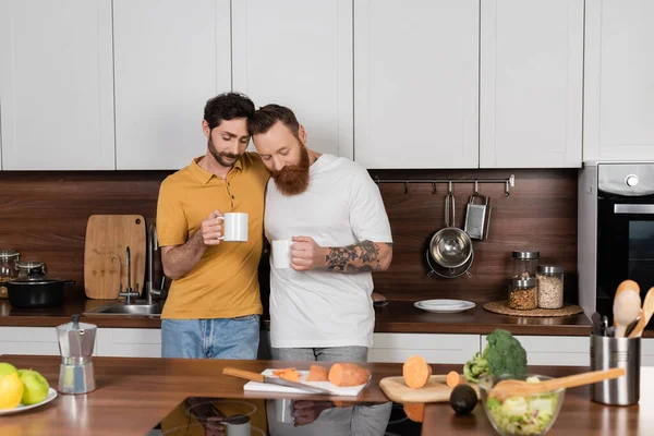 Gay couple holding cups of coffee near vegetables in kitchen — Stock Photo