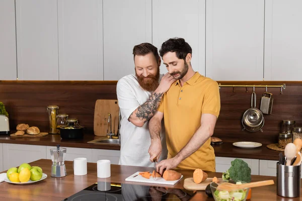 Улыбающийся гей обнимает партнера и готовит дома на кухне — стоковое фото