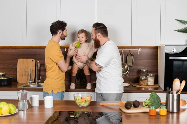 Cheerful gay couple holding toddler daughter with apple in kitchen — Photo de stock