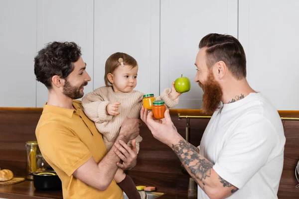 Gay couple holding toddler daughter and baby food in kitchen — Foto stock