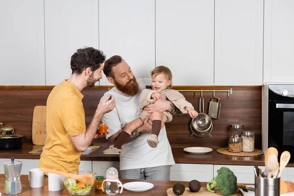 Gay parents holding baby food near displeased daughter in kitchen — Stockfoto