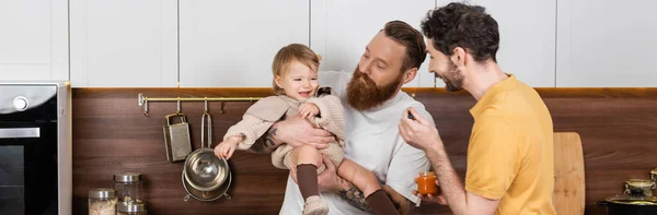 Smiling gay couple holding baby food near toddler daughter in kitchen, banner — Photo de stock