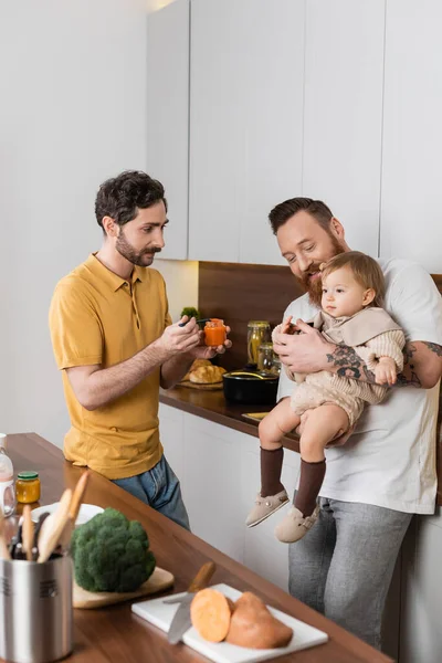Smiling gay man holding toddler daughter near husband with baby food in kitchen — Stock Photo