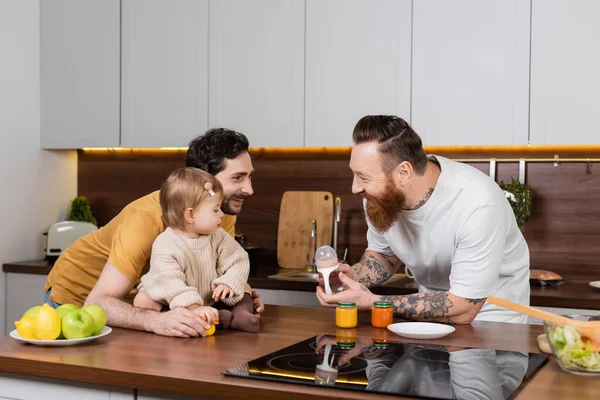 Gay casal segurando bebê garrafa perto criança filha no cozinha — Fotografia de Stock
