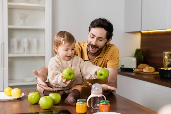 Father looking at baby daughter holding apples near baby food and bottle in kitchen — Stockfoto