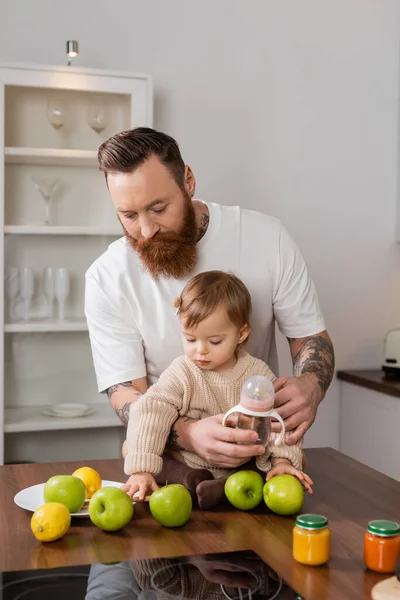Татуйований тато тримає дитячу пляшку біля дочки та фрукти на кухні — стокове фото