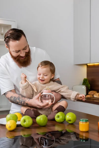 Бородатый мужчина держит детскую бутылочку рядом с дочерью с ложкой и фруктами на кухне — стоковое фото