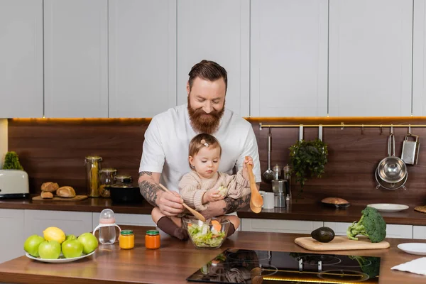 Татуйований батько посміхається і змішує салат біля дочки малюка на кухні — стокове фото