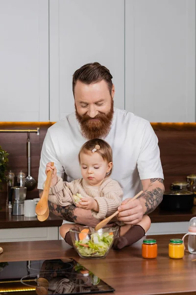 Smiling father cooking salad near daughter and baby food in kitchen — Foto stock