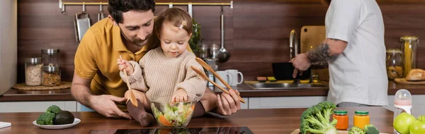 Homosexual parent holding daughter with spoon and fresh salad in kitchen, banner — Stock Photo